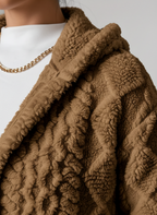 Close-up of the bouclé-like raised zigzag and diamond relief texture on the camel brown hooded cardigan, with the folded hood collar visible