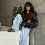 Seated model wearing the dark brown oversized moto jacket with grommet belt tied at the waist, paired with wide-leg jeans against a concrete backdrop