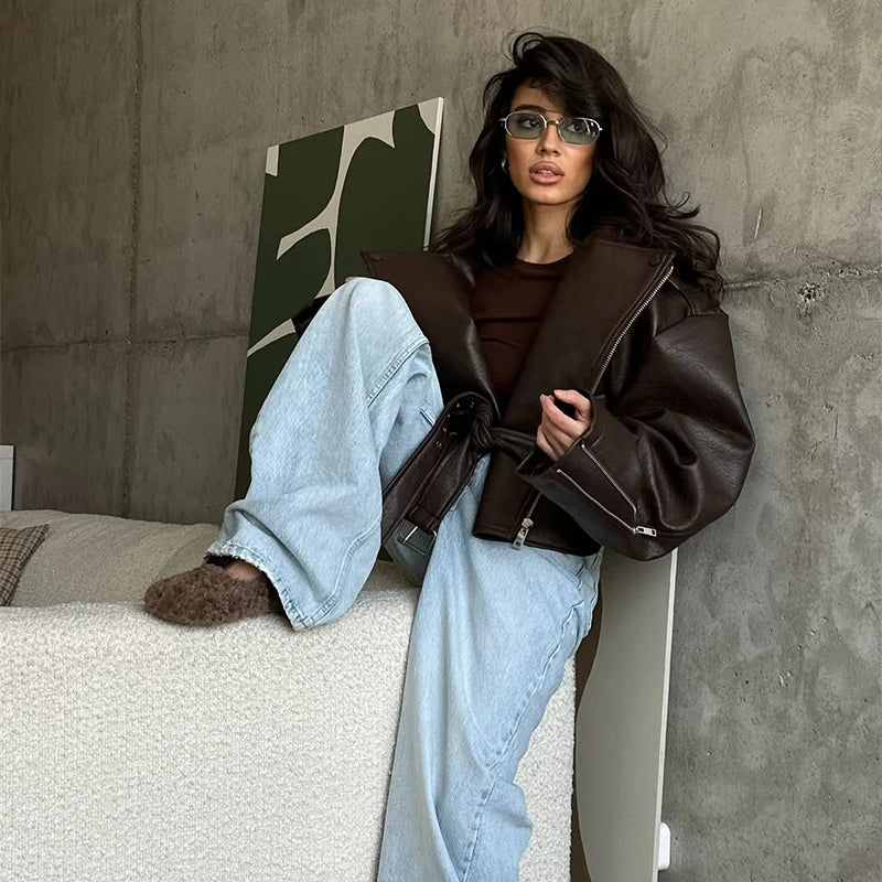 Seated model wearing the dark brown oversized moto jacket with grommet belt tied at the waist, paired with wide-leg jeans against a concrete backdrop
