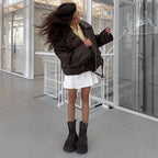Woman in motion wearing the dark brown oversized moto jacket open over a white mini skirt and chunky boots, showing the dropped-shoulder silhouette