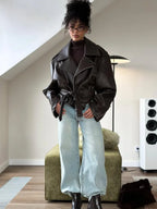 Full-length view of a woman in a dark brown oversized moto jacket belted at the waist, worn over a turtleneck and wide-leg jeans with black boots