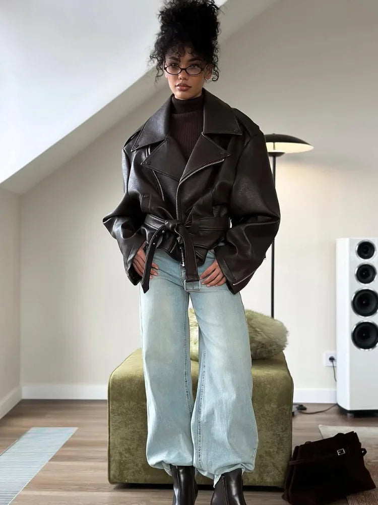 Full-length view of a woman in a dark brown oversized moto jacket belted at the waist, worn over a turtleneck and wide-leg jeans with black boots